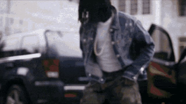 a man in a denim jacket is standing in front of a car .