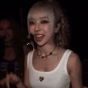 a woman wearing a white tank top and a necklace is standing in front of a microphone in a dark room .