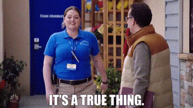 a woman in a blue shirt is talking to a man in a brown vest while holding a book .