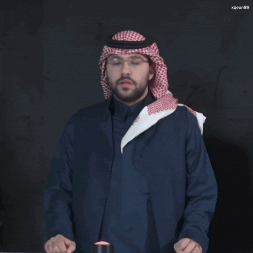 a man wearing a keffiyeh and glasses is standing in front of a black background with xqeon89 written on it