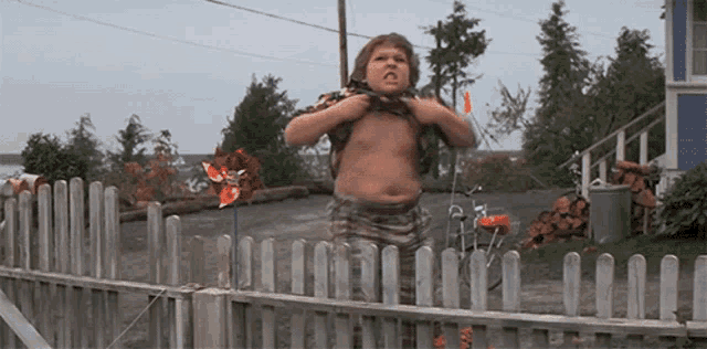 a shirtless boy stands in front of a wooden fence
