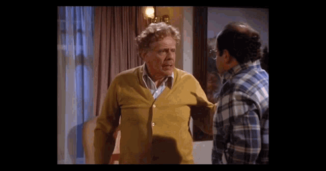 an older man in a yellow cardigan talks to another man in a plaid shirt