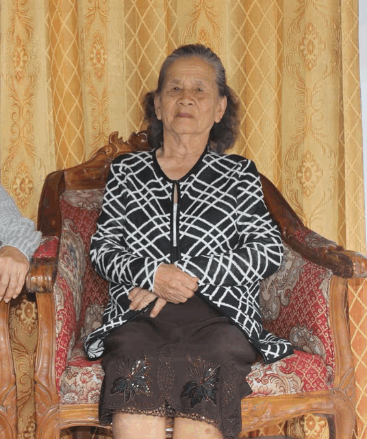 an elderly woman sits in a chair wearing a black and white jacket