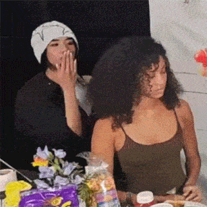 two women are sitting at a table with food and one is covering her mouth with her hand .