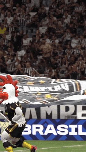 a rooster mascot is running on a soccer field in front of a brasil banner