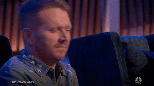a man in a denim shirt is sitting in a blue chair with his eyes closed and a nbc logo in the background