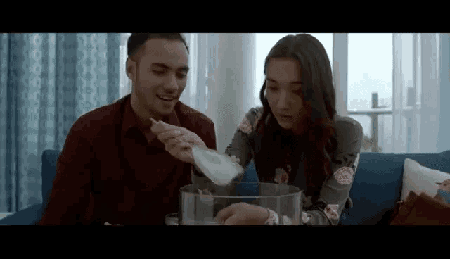 a man and a woman are sitting on a couch looking at a bowl of food .