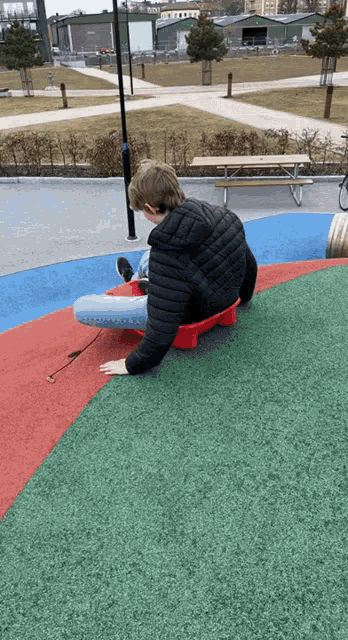 a boy in a black jacket is sitting on a red stool in a park