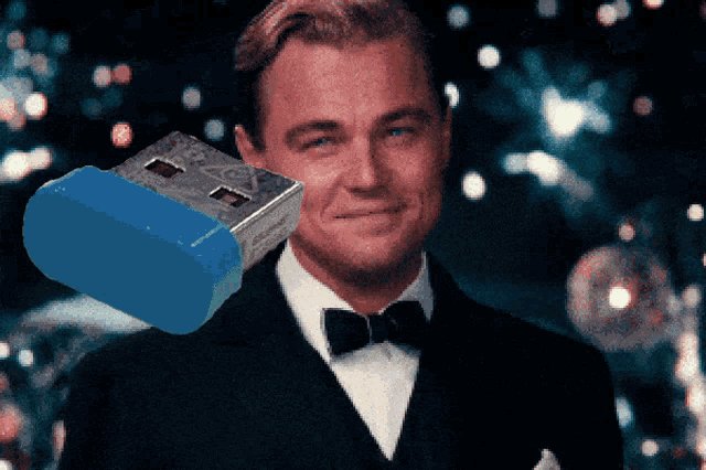 a man in a tuxedo and bow tie is smiling with a blue flash drive in front of him