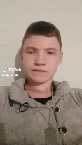 a young man is wearing a grey jacket and a chain around his neck while looking at the camera ..