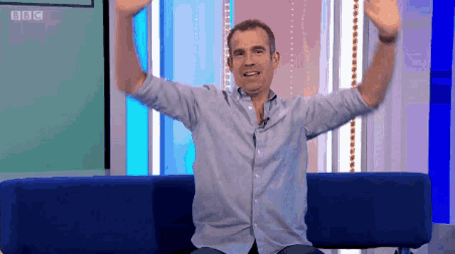a man is sitting on a blue couch with his arms in the air in front of a wall that says bbc