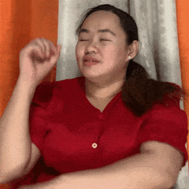 a woman in a red shirt is sitting in front of an orange curtain with her eyes closed