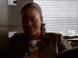 a woman is sitting in front of a window with blinds and making a funny face .