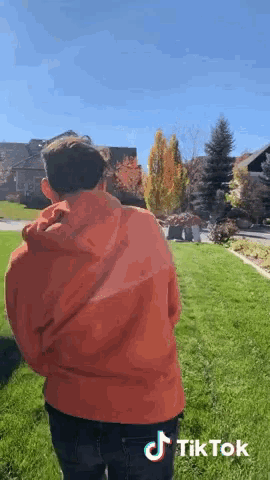 a man in an orange hoodie is standing in a grassy field with a tiktok logo on the bottom right corner .