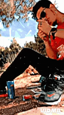a man wearing sunglasses sits on the ground smoking a cigarette and drinking soda