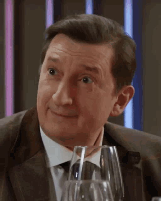a man in a suit and tie is smiling while sitting at a table with wine glasses