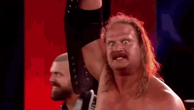 a man with long hair and a mustache is making a funny face while holding his arm in the air .