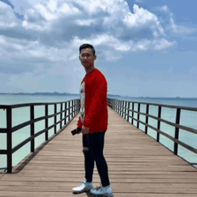 a man in a red sweater is standing on a wooden pier overlooking a body of water .