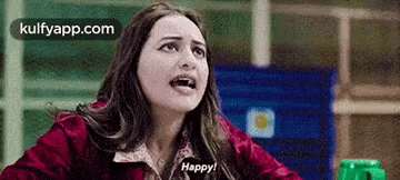 a woman in a red jacket is sitting at a table with her mouth open and saying happy .