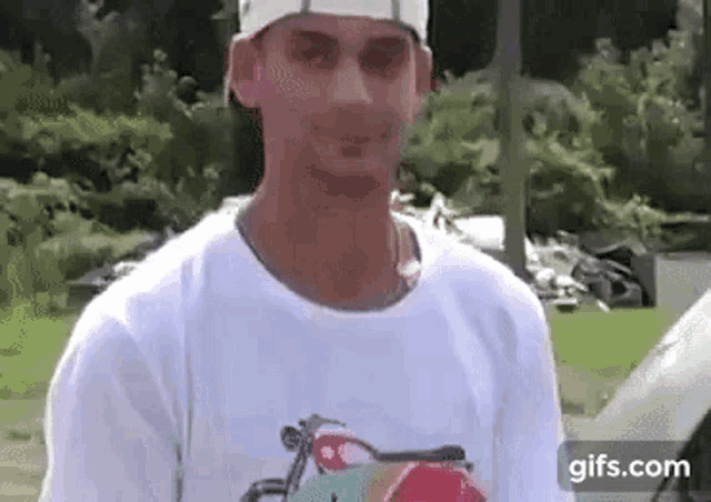 a man wearing a white hat and a white t-shirt is standing in front of a car and looking at the camera .