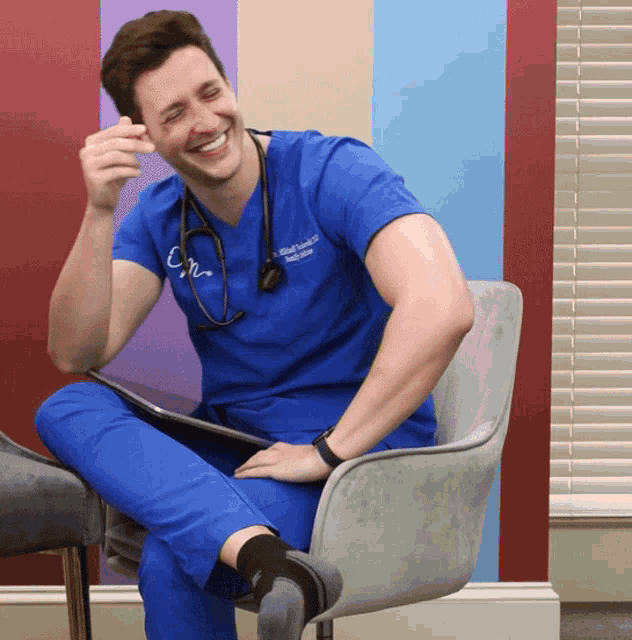 a man in a blue scrub top with a stethoscope around his neck is sitting on a chair