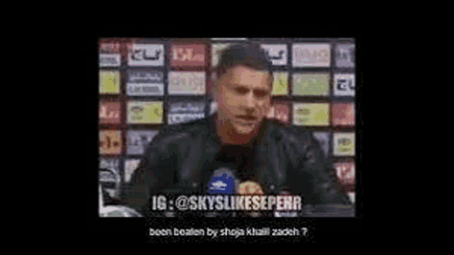 a man is sitting at a table talking into a microphone in front of a soccer field .