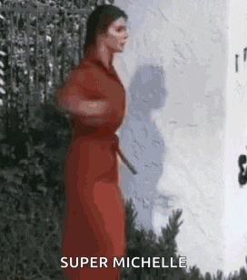 a woman in a red dress is standing in front of a white wall with the words `` super michelle '' written on it .