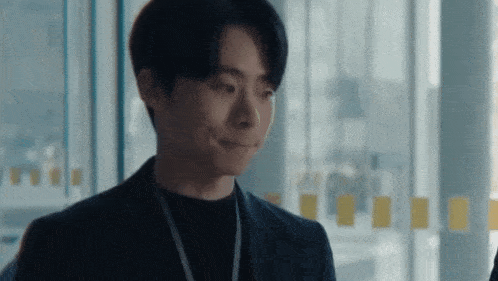 a man in a suit and black sweater is making a funny face while standing in front of a window .