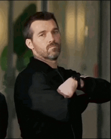 a man with a beard wearing a black jacket and a watch is looking at his watch .