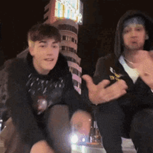 two young men are sitting next to each other in front of a building with a clock on it