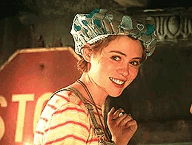 the woman is wearing a hat and smiling in front of a stop sign .