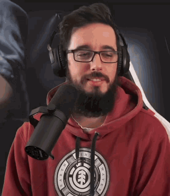 a man with a beard wearing headphones and a red hoodie is standing in front of a microphone and smiling .