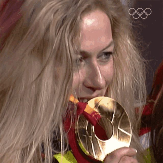 a woman is holding a gold medal in her mouth with the olympic rings in the background