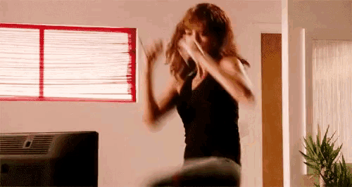 a woman in a black tank top is dancing in a living room in front of a window .