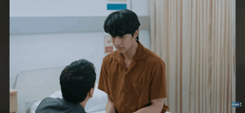 a man in a brown shirt is sitting next to another man in a hospital room .