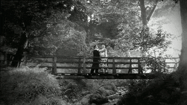 a black and white photo of two people standing on a bridge .