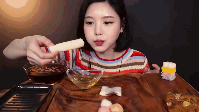 a woman is sitting at a table with a bowl of syrup and a stick of food
