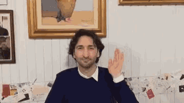 a man in a blue sweater is waving his hand in front of a painting on a wall .