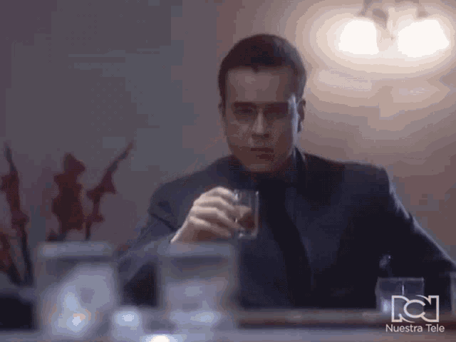 a man in a suit and tie is sitting at a table drinking from a glass with nuestra tele written on the bottom