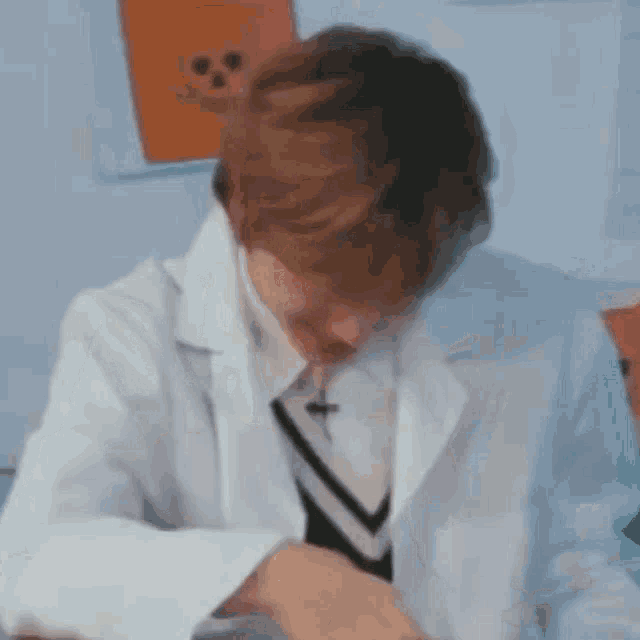 a man in a white coat and tie is sitting at a table with his hands on his face .