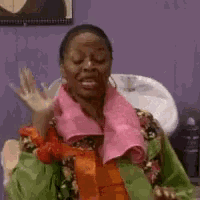 a woman with a pink towel around her neck is sitting in front of a sink and waving .