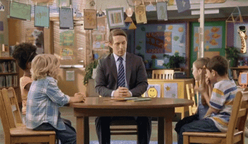a man in a suit sits at a table with children in a classroom