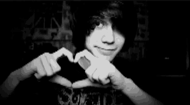 a black and white photo of a young man with long hair making a heart shape with his hands .