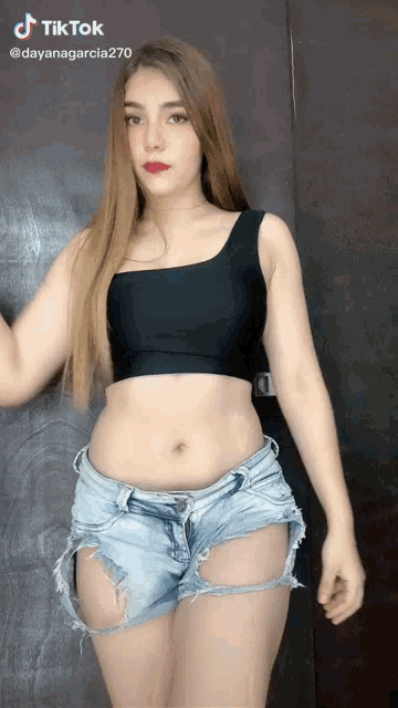 a woman in a black crop top and denim shorts stands in front of a brown door