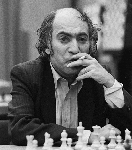 a man in a suit is sitting at a table with chess pieces and smoking a cigarette .