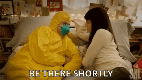 a man in a yellow protective suit is sitting on a bed with a woman and says " be there shortly "