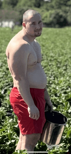 a shirtless man in red shorts is holding a metal bucket in a field