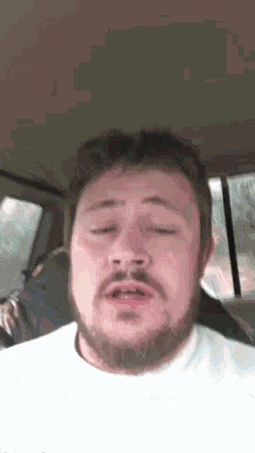 a man with a beard is sitting in a car with his eyes closed and making a funny face .