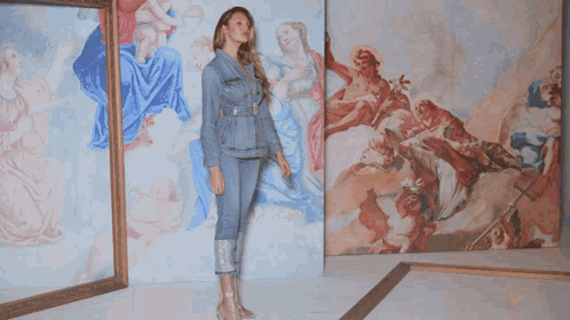 a woman in a denim jacket stands in front of a wall of paintings