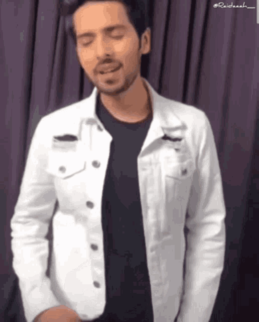 a man wearing a white jacket and black shirt is standing in front of a purple curtain